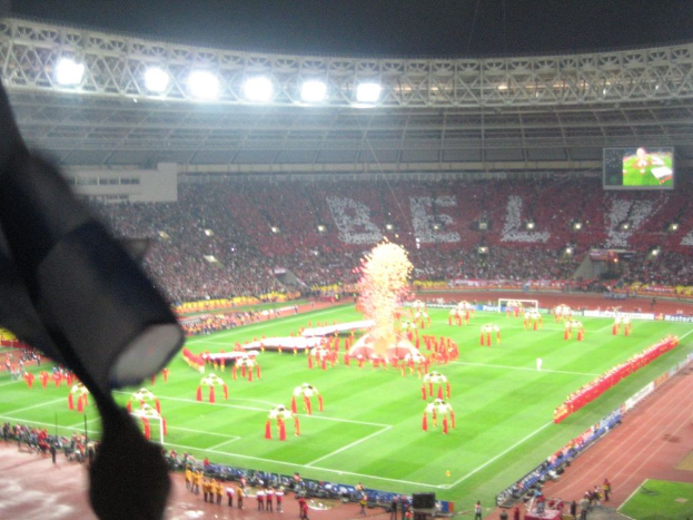 Ein Stadion mit Flutlicht, Eisengittern, Zuschauern, stehenden Personen, Werbetafeln, Bildschirmen, Sicherheitskameras, Fahnen und Dekorationen.