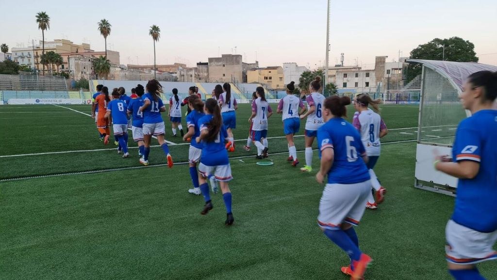 Eine Gruppe von Frauen, die auf einem städtischen Feld Fußball spielen, mit einem Tor und Netz im Hintergrund.