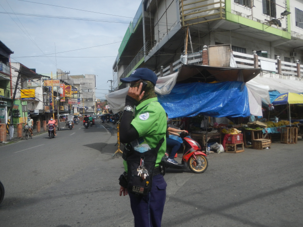 Polizist in grüner und blauer Uniform mit blauer Mütze auf einer Straße stehend, mit einem Handy telefonierend, mit Fahrzeugen, Marktständen, einem Gebäude und bewölktem Himmel im Hintergrund.