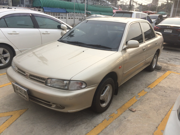 Ein goldfarbener Wagen auf einem Parkplatz neben anderen Fahrzeugen geparkt, mit Gebäuden, Strommasten, Drähten, Bäumen und einem klaren blauen Himmel im Hintergrund.