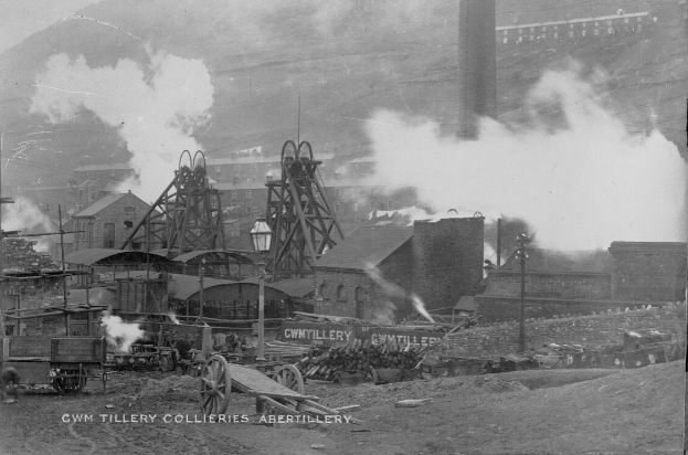 Schwarzes und weißes Foto einer rauchenden Fabrik, umgeben von Gebäuden, Pfählen, Fahrzeugen, Karren und anderen Gegenständen, mit Text unten.