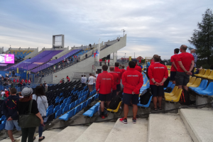 Eine Gruppe von Menschen, einige sitzen und andere stehen auf einer Treppe, versammelt vor einem Stadion mit einem Baum auf der rechten Seite, einem Bildschirm auf der linken Seite, Laternenmasten und einem klaren blauen Himmel im Hintergrund.