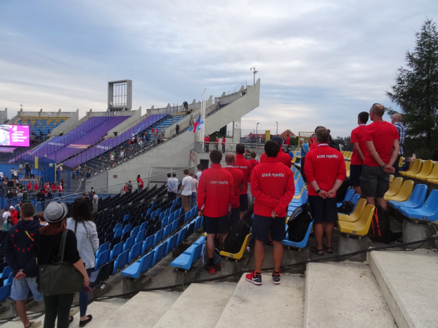 Eine Gruppe von Menschen, einige sitzen und andere stehen auf einer Treppe, versammelt vor einem Stadion mit einem Baum auf der rechten Seite, einem Bildschirm auf der linken Seite, Laternenmasten und einem klaren blauen Himmel im Hintergrund.