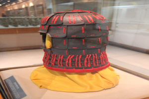 Ein gelber, mit Stoff bedeckter Helm, der in einer Glasvitrine ausgestellt ist, mit einer Texttafel links daneben und Deckenbeleuchtung im Hintergrund.