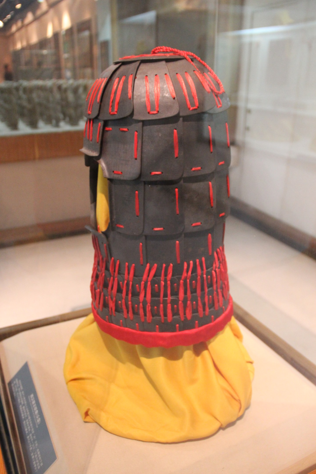 Ein gelber, mit Stoff bedeckter Helm, der in einer Glasvitrine ausgestellt ist, mit einer Texttafel links daneben und Deckenbeleuchtung im Hintergrund.