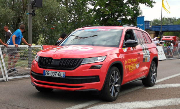 Roter Wagen fährt auf einer Straße neben einer Menge, einige tragen Kappen, mit Maschendrahtzaun links, einem Verkehrssignalmast, Bäumen, einer Brücke, einem Fahnenmast und bewölktem Himmel im Hintergrund.