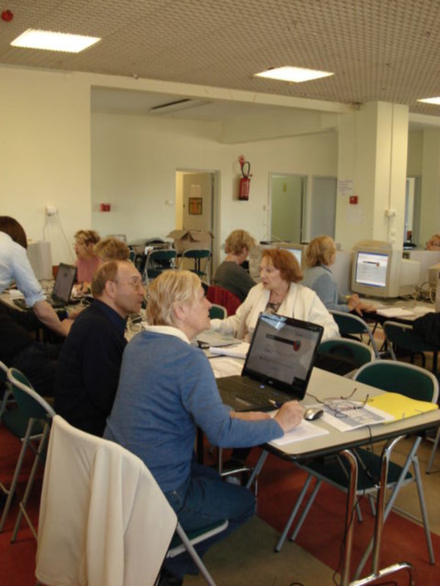 Ein Raum mit Menschen, die auf Stühlen sitzen und auf Laptops arbeiten, ein Tisch mit leeren Stühlen in der Mitte, eine Decke mit Lampen, eine Wand mit einem Feuerlöscher und zwei Türen.