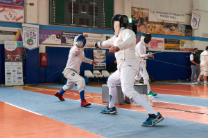 Eine Gruppe von Menschen in Helmen und mit Schwertern ficht in einer Turnhalle mit Bannern, einem Bildschirm und verstreuten Stühlen im Hintergrund.