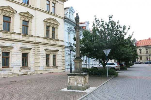 Bronzestatuette einer stehenden Figur auf einem Sockel in einem Stadtplatz, umgeben von Gebäuden, Straßeninfrastruktur, Fahrzeugen, Vegetation und einem klaren Himmel.