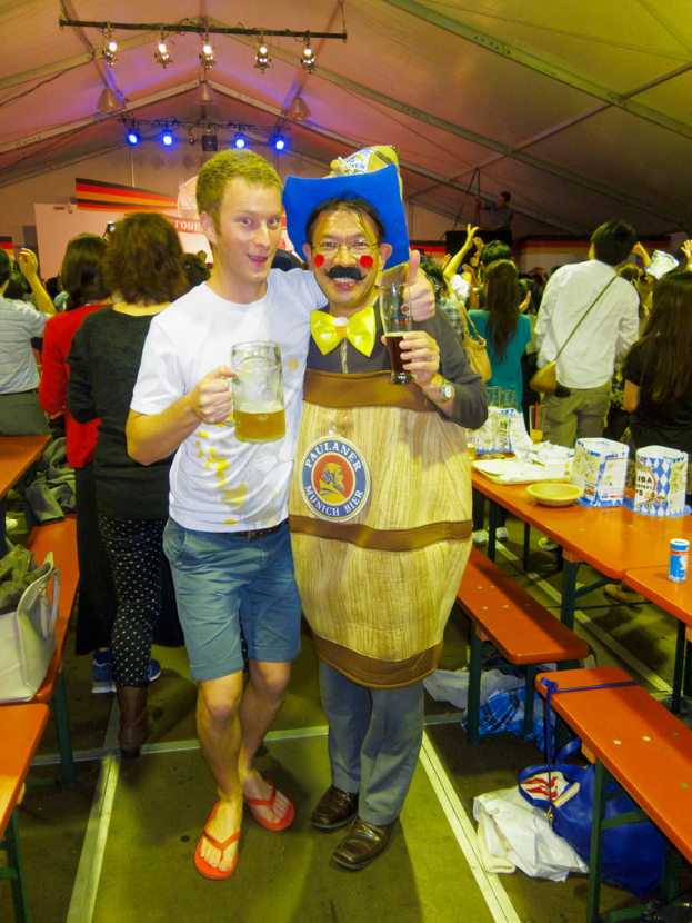 Zwei Personen stehen zusammen, eine hält ein Kind und ein Bier, die andere trägt ein Kostüm, mit einem Tisch voller Essen in der Nähe und einer Gruppe von Menschen dahinter.