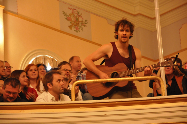 Eine Gruppe von Menschen, die Musik hören und genießen, mit einem Mann in der Mitte in einem ärmellosen Hemd, der singt und eine Gitarre spielt, vor einer braunen Wand mit einem Bild oben.