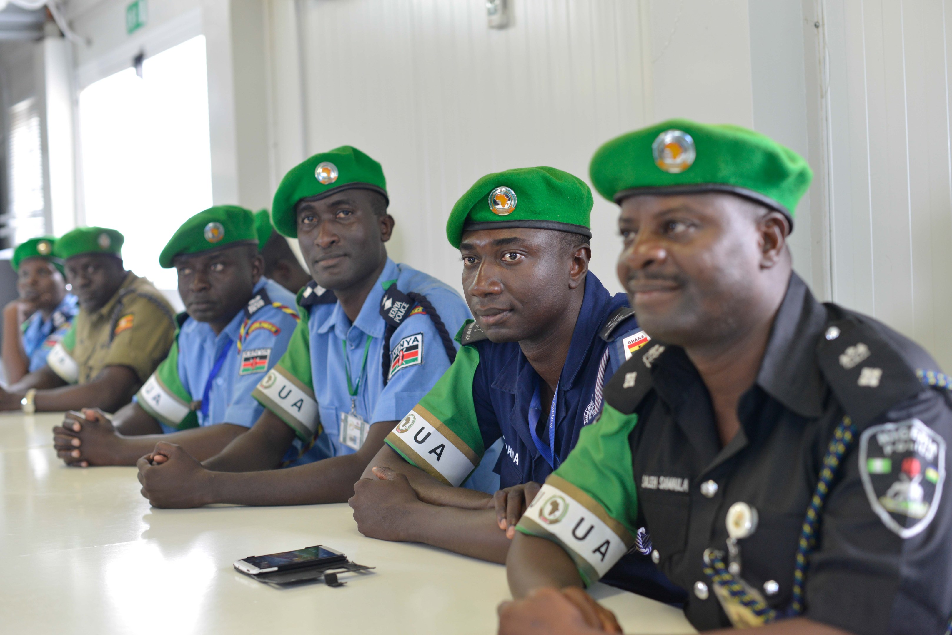 Gruppe von Polizeibeamten bei einer Schulung an einem Tisch sitzend, mit Mobiltelefonen und anderen Gegenständen auf dem Tisch, in einem Raum mit Fenstern und Deckenleuchten.