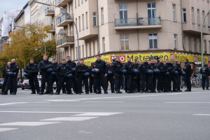 Eine Gruppe von Polizisten in schwarzen Uniformen mit blauen Helmen, die auf einer Straße mit Laternen, Bäumen und glasverkleideten Gebäuden unter einem klaren blauen Himmel stehen.