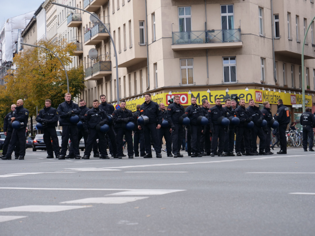 Eine Gruppe von Polizisten in schwarzen Uniformen mit blauen Helmen, die auf einer Straße mit Laternen, Bäumen und glasverkleideten Gebäuden unter einem klaren blauen Himmel stehen.