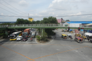 Eine belebte Stadtstraße mit Autos, Bussen, Fußgängern auf einem Gehweg, einer Brücke mit Geländern, Ampeln, Bäumen, Gebäuden, Strommasten mit Drähten und einem bewölkten Himmel.