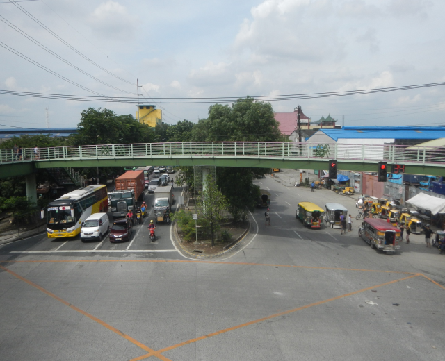 Eine belebte Stadtstraße mit Autos, Bussen, Fußgängern auf einem Gehweg, einer Brücke mit Geländern, Ampeln, Bäumen, Gebäuden, Strommasten mit Drähten und einem bewölkten Himmel.