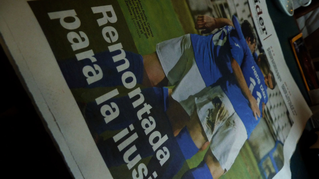 Eine Zeitung auf einem Tisch zeigt drei MÃ¤nner in blauen T-Shirts, mÃ¶glicherweise beim Fussballspielen, mit Text unten auf dem Bild.