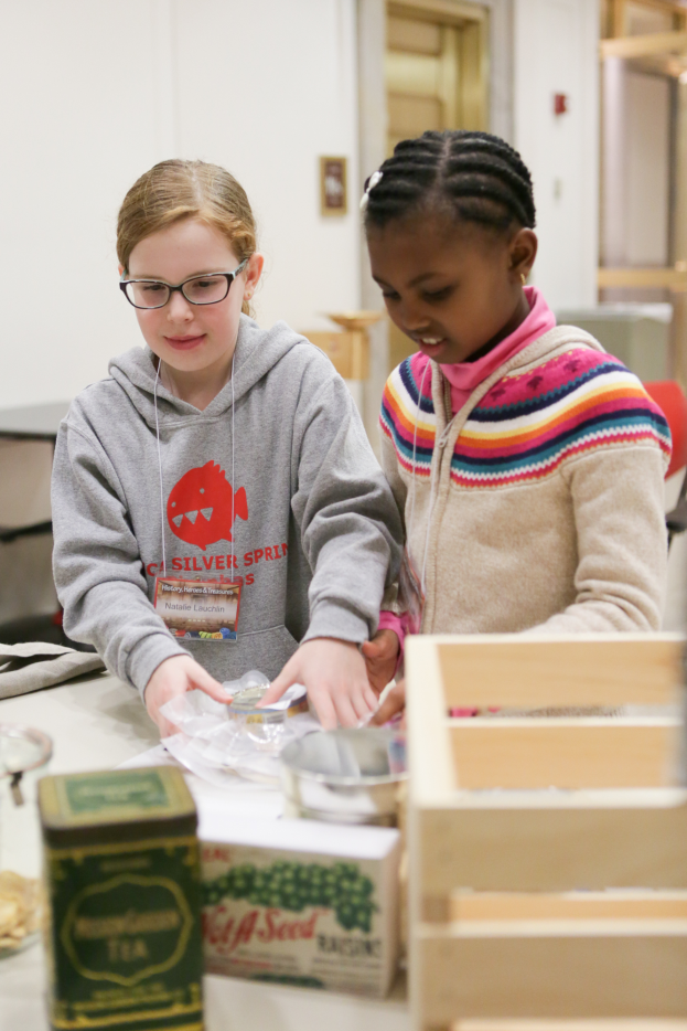 Zwei junge Mädchen arbeiten an einem Projekt an einem Tisch mit Kisten, Schüsseln und anderen Gegenständen in einem Klassenzimmer, mit einer Wand und einer Tür im Hintergrund.