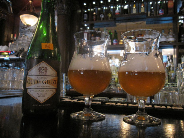 Eine Flasche mit einem Logo und zwei Weingläser auf einem Tisch, mit vielen Gläsern, Flaschen und Gestellen im Hintergrund.