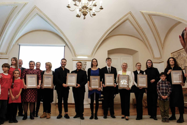 Eine Gruppe von Menschen steht nebeneinander in einem Raum, jeder hält ein Zertifikat in der Hand, mit einer Hand auf der rechten Seite, einer Wand, einer Tür, einem Bildschirm und einem Kronleuchter im Hintergrund, wahrscheinlich bei einer Preisverleihung für das akademische Jahr 2018-2019.