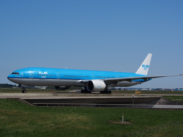 KLM Boeing 777-300ER Flugzeug auf der Rollbahn am Amsterdam Airport, mit Gras, einem Zaun mit Pfählen, Gebäuden, Bäumen und einem klaren blauen Himmel im Hintergrund.