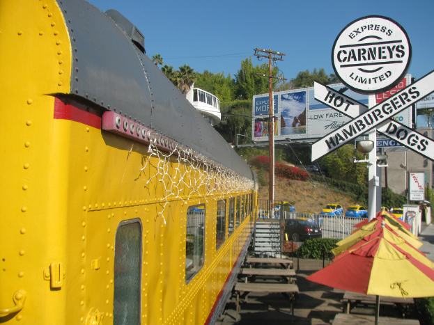 Ein mobiler Imbiss ist in der Nähe von Bänken und Treppen geparkt, mit Schirmen, Pflanzen und einem Geländer in der Nähe. Fahrzeuge, ein Pfahl, Schilder und eine Absperrung sind ebenfalls zu sehen, sowie ein Gebäude, Bäume und verschiedene Objekte unter einem klaren Himmel.