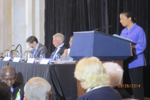 Eine Frau steht an einem Tisch und spricht während einer Konferenz, mit drei sitzenden Männern neben ihr an einem anderen Tisch, der Gläser, Bücher und Namensschilder zeigt, während einige Menschen vor den Tischen arbeiten und ein Vorhang und eine Wand im Hintergrund zu sehen sind.