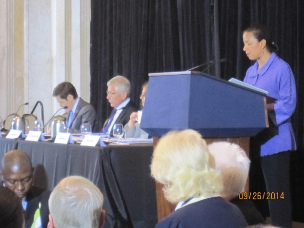 Eine Frau steht an einem Tisch und spricht während einer Konferenz, mit drei sitzenden Männern neben ihr an einem anderen Tisch, der Gläser, Bücher und Namensschilder zeigt, während einige Menschen vor den Tischen arbeiten und ein Vorhang und eine Wand im Hintergrund zu sehen sind.