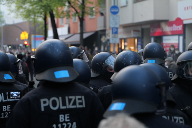 Polizisten in schwarzen Uniformen und Helmen stehen vor einer Menge, mit unscharfen Gebäuden, Bäumen und Verkehrszeichen im Hintergrund.