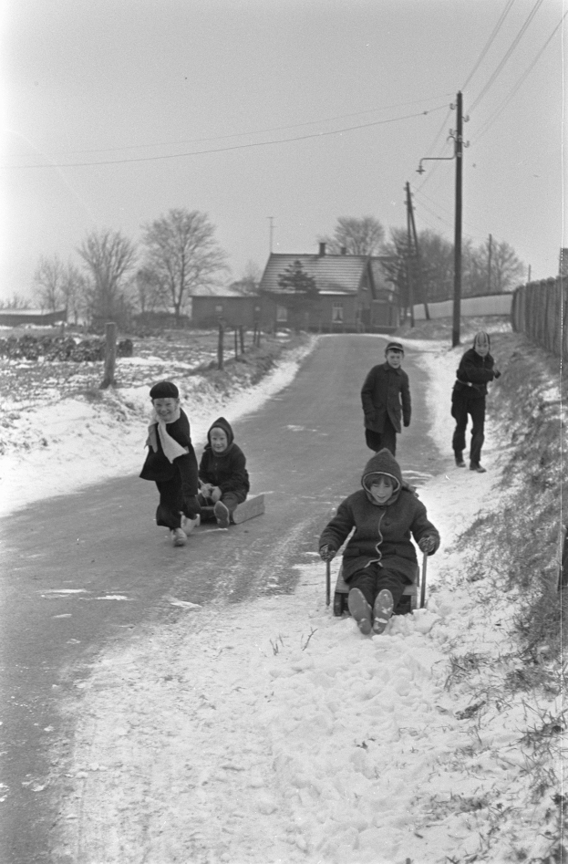 Eine Gruppe von Kindern in Mänteln und Mützen fährt auf einer verschneiten Straße Schlitten, mit Häusern, Bäumen, Pfählen, Drähten und einem Zaun im Hintergrund, unter einem klaren Himmel, dargestellt in Schwarz-Weiß.