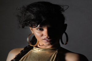 Eine Frau in einem goldenen Kleid und großen Ohrringen steht selbstbewusst vor einer Wand, ihr glatter Bob und natürliches Make-up unterstreichen ihren entschlossenen Gesichtsausdruck, während sie direkt in die Kamera schaut.