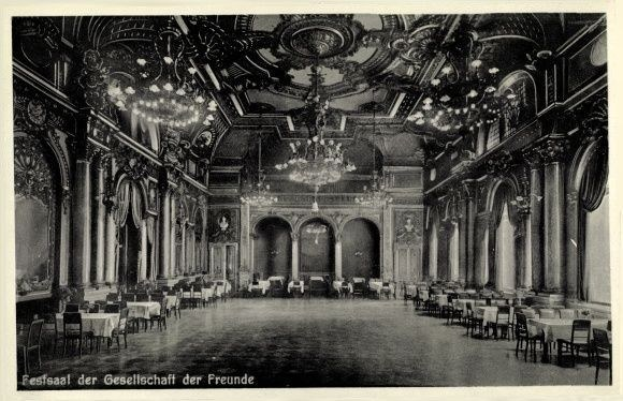 Schwarzes und weißes Foto eines großen Raumes mit Tischen, Stühlen, Säulen, Bögen und Deckenleuchtern, beschriftet mit "Fest der Gesellschaft der Freunde, Berlin" unten.