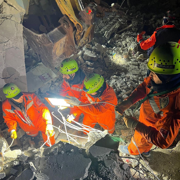 Eine Gruppe von Menschen in orangefarbenen Uniformen und grünen Helmen bei der Arbeit an Schutt von einem eingestürzten Gebäude, mit einem Kran im Hintergrund.