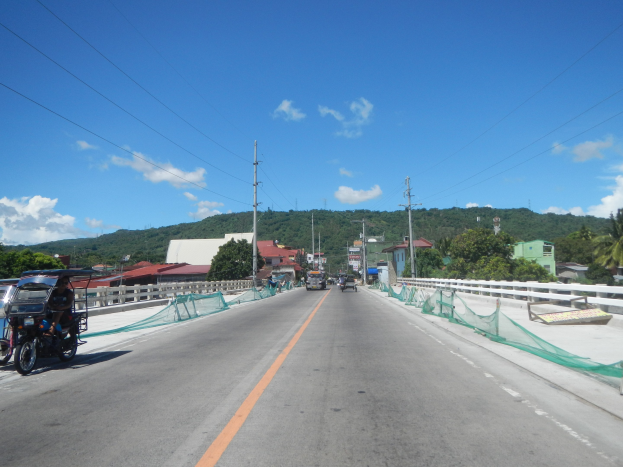 Autobahn mit fahrenden Fahrzeugen, ein geparktes Motorrad auf dem Seitenstreifen, Geländer, Strommäste mit Drähten, Bäume, Gebäude, ferne Hügel und ein bewölkter Himmel.