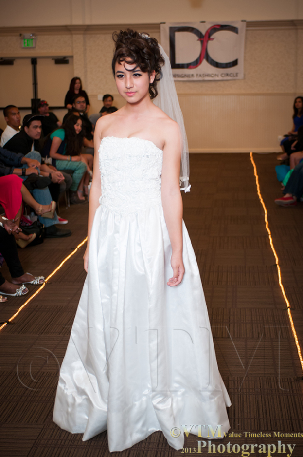 Eine Frau in einem weißen Kleid lächelt und posiert für ein Foto vor einem weißen Banner mit Text darauf, während Menschen auf Stühlen auf beiden Seiten von ihr sitzen und ein brauner Boden unten sichtbar ist.