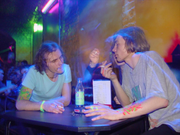 Ein Tisch mit einer Flasche, einer Speisekarte und einem Aschenbecher in der Mitte einer Indoor-Einrichtung, mit zwei Männern und einer Frau, die Zigaretten rauchen.