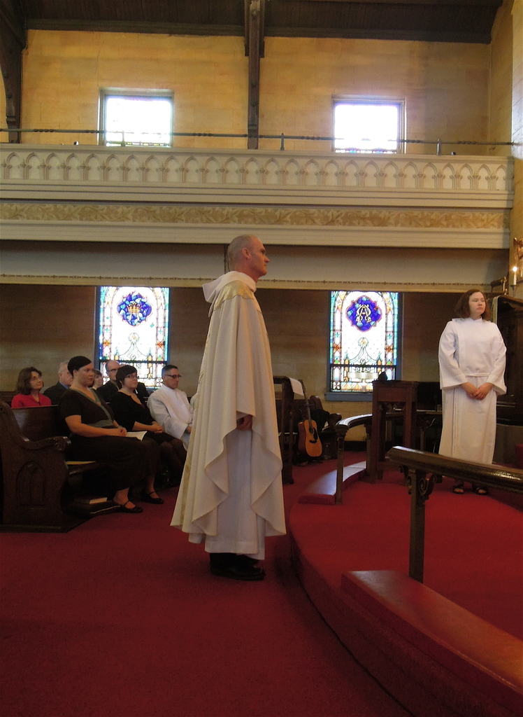 Eine Frau und ein Mann in weißen Kleidern stehen in einer Kirche, mit einem roten Teppich zu ihren Füßen, hölzernen Geländern, Kerzen, Bänken mit sitzenden Menschen und Musikinstrumenten im Hintergrund.
